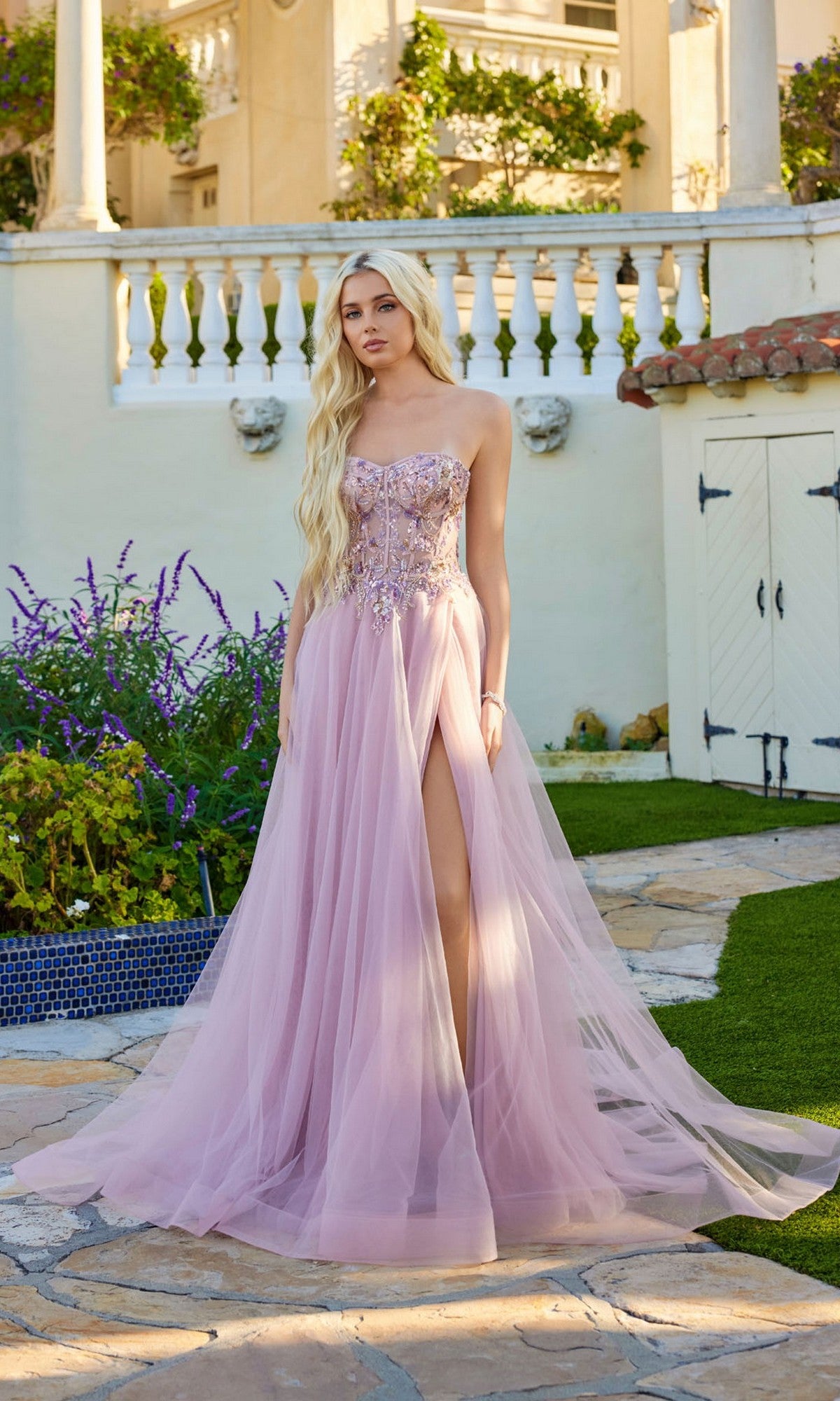 A woman wears the Juliet JT2640G long prom dress, featuring a strapless lavender tulle design with a sheer skirt, thigh-high slit, and lace-up back, standing on a stone path in a garden.